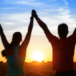 Successful couple of young athletes raising arms to golden summer sunset sky after training. Fitness man and woman with arms up celebrating sport goals after exercising in countryside field.